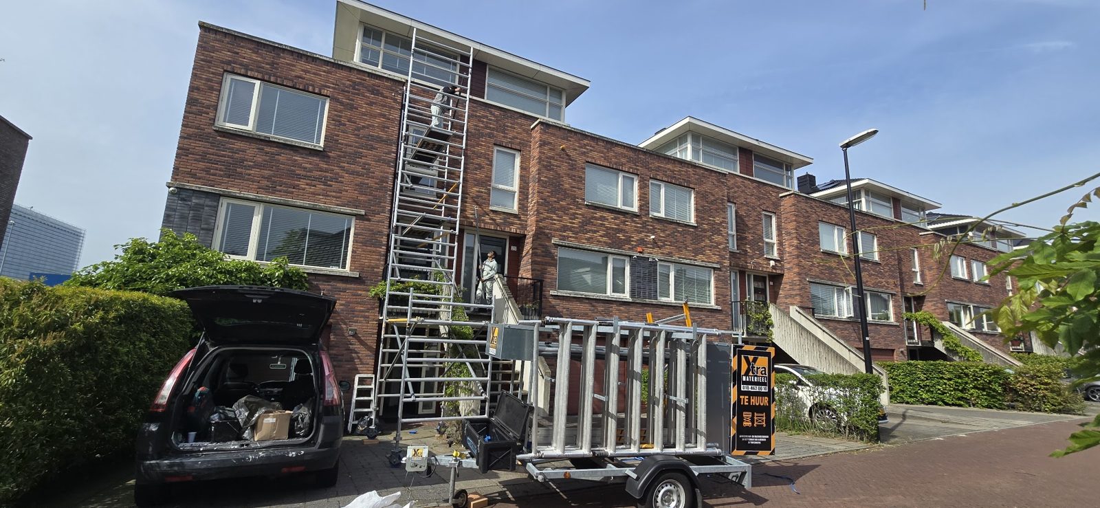 Rolsteiger tegen een bakstenen rijtjeshuis, met een aanhanger met steigermateriaal ervoor